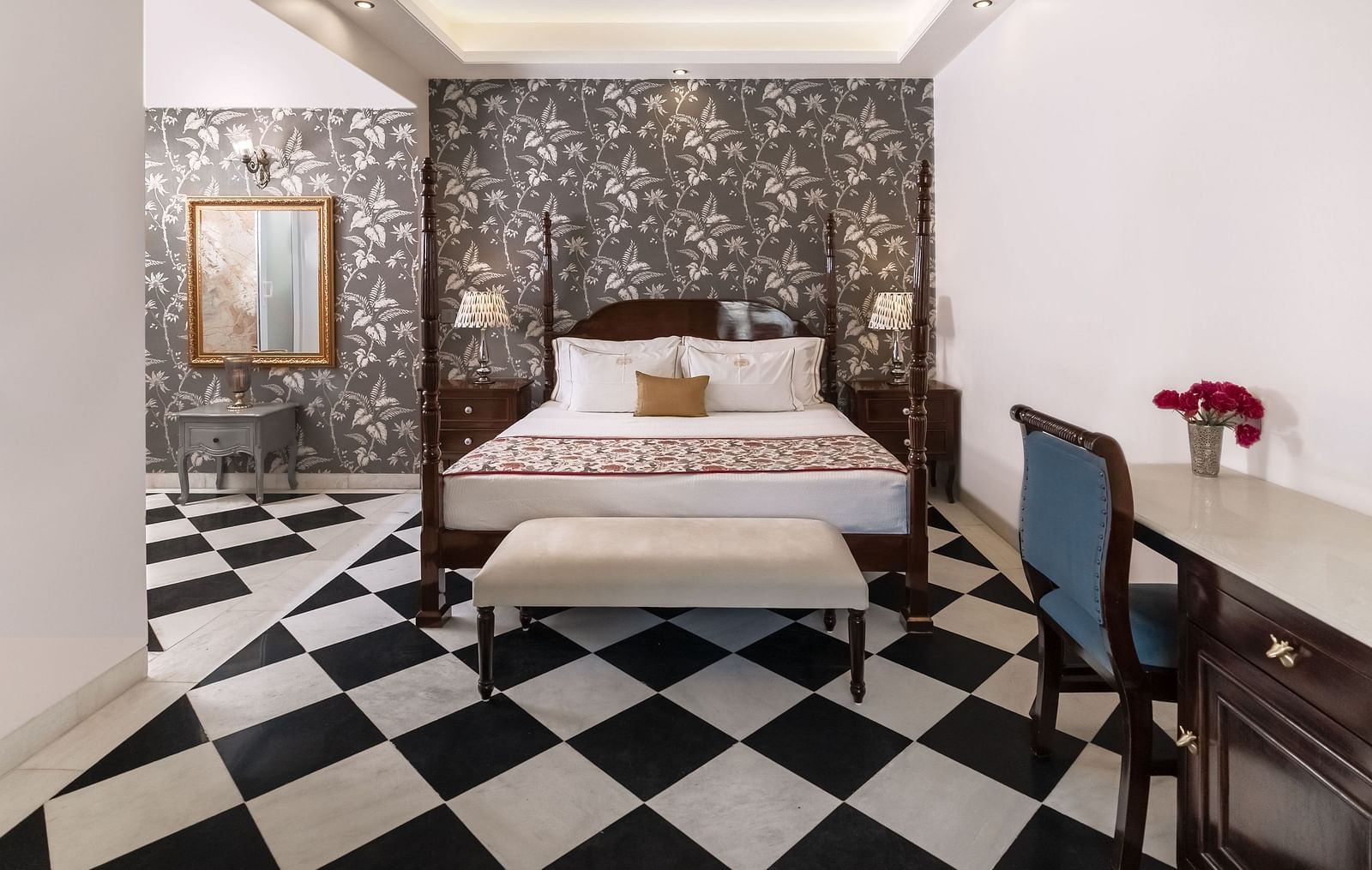 A bedroom with a four-poster bed, a patterned wallpapered wall, a chequered black and white tiled floor, and a writing desk with a blue chair | Dileep  Kothi, Jaipur